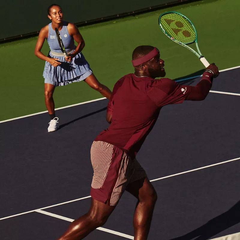 Pro Tennis Players and lululemon Ambassadors Frances Tiafoe and Leylah Fernandez wear the newest tennis gear as Frances reaches for a shot at the net and Leylah covers the baseline behind them. 